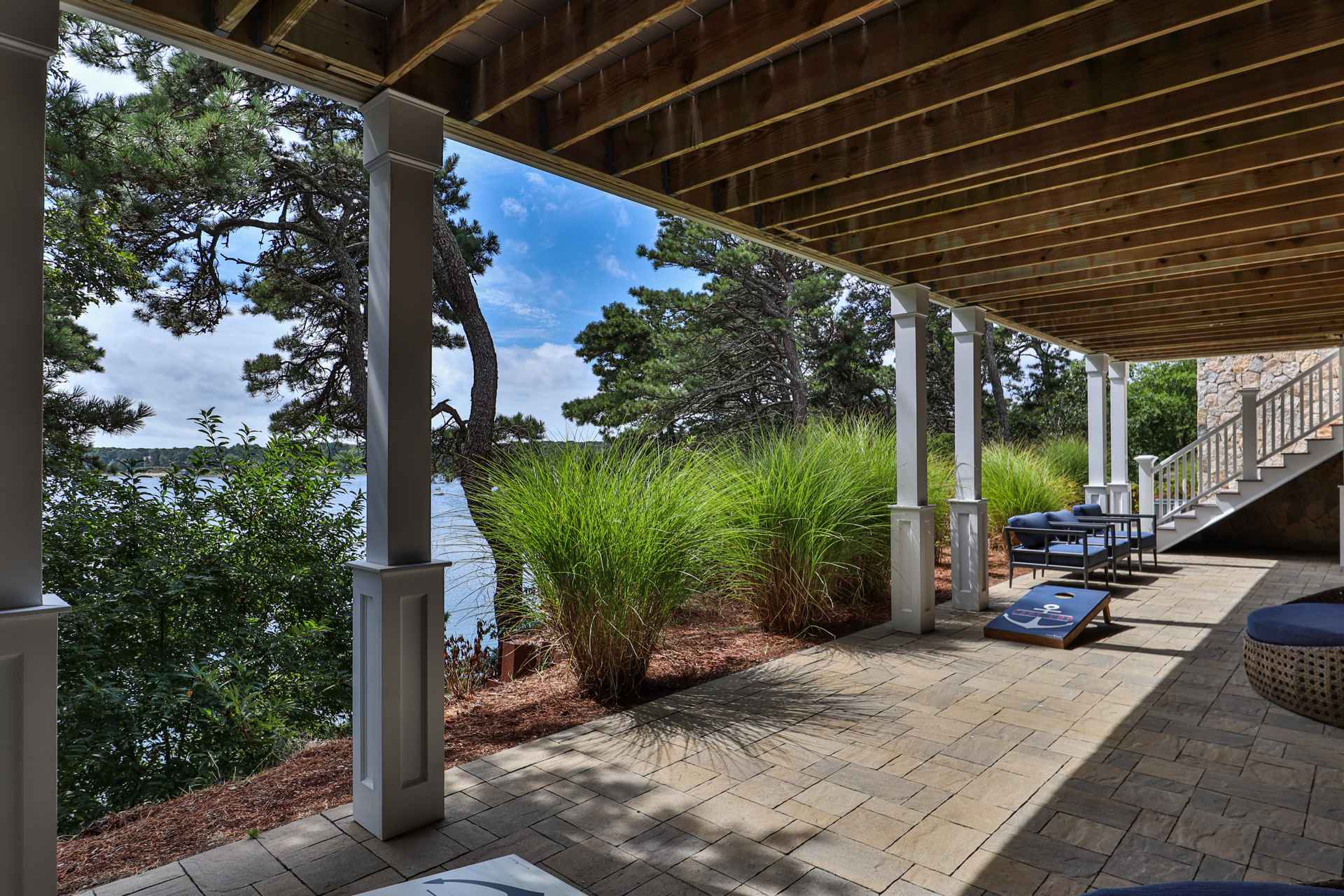This image showcases a covered patio area with a view of a lake or ocean. The patio features a stone paver floor, white support columns, and comfortable outdoor seating. Lush greenery and trees surround the area, creating a serene and inviting outdoor living space, perfect for relaxation and entertaining.