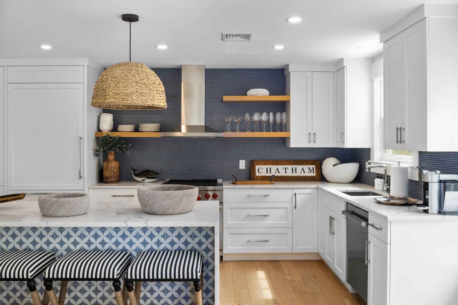 This is a bright and modern kitchen featuring white cabinetry, stainless steel appliances, and a stylish island with a blue patterned base and white countertop. The backsplash is a dark blue tile, complemented by wooden shelves and a woven pendant light. The perspective is a wide shot, capturing the entire kitchen space.