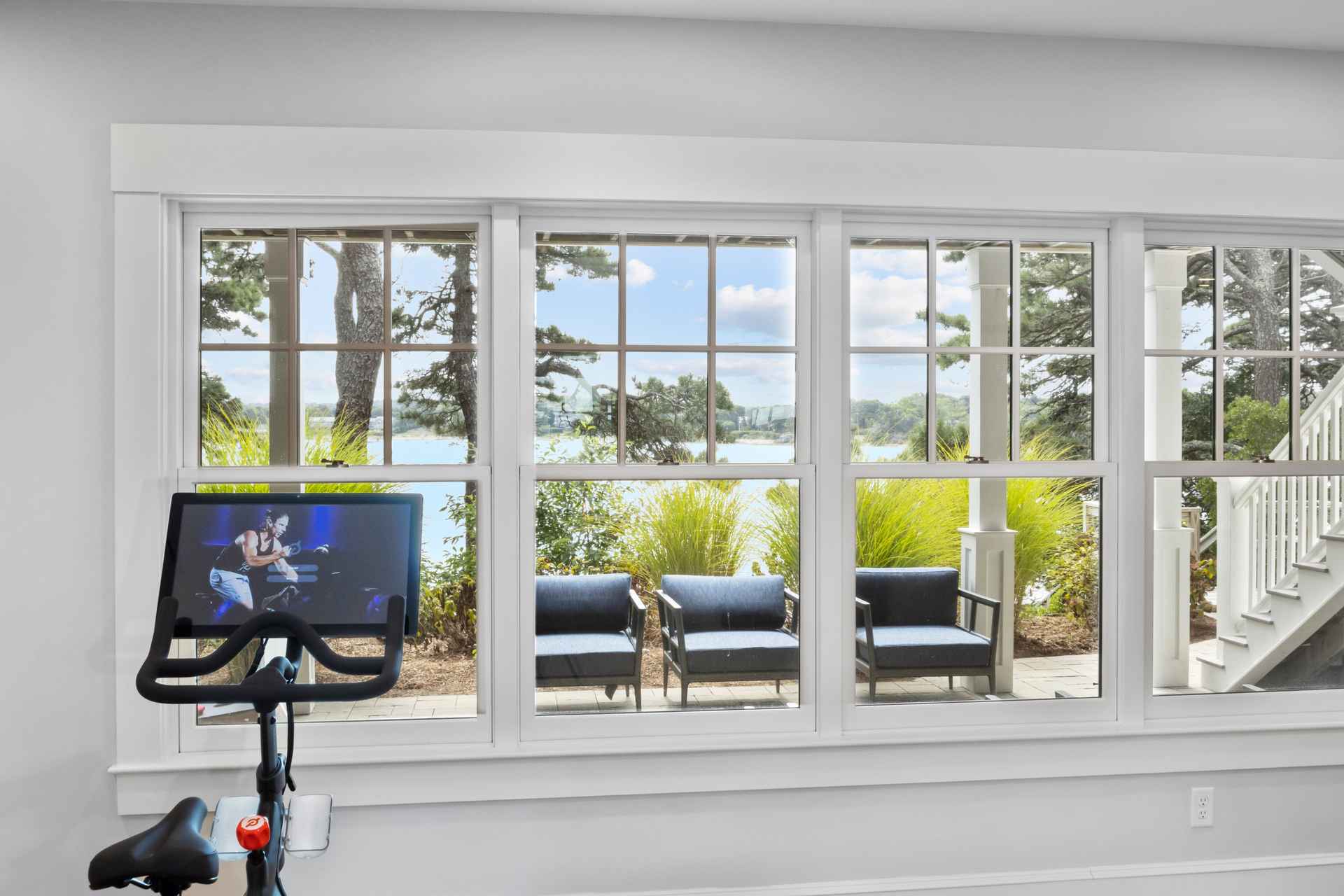 This image showcases a home gym area with a stationary bike positioned in front of a large window offering a scenic view of a lake and outdoor seating. The room is bright and airy, with white trim and walls, creating a clean and inviting workout space. The view outside features a patio with chairs and lush greenery, enhancing the appeal of the indoor-outdoor connection.