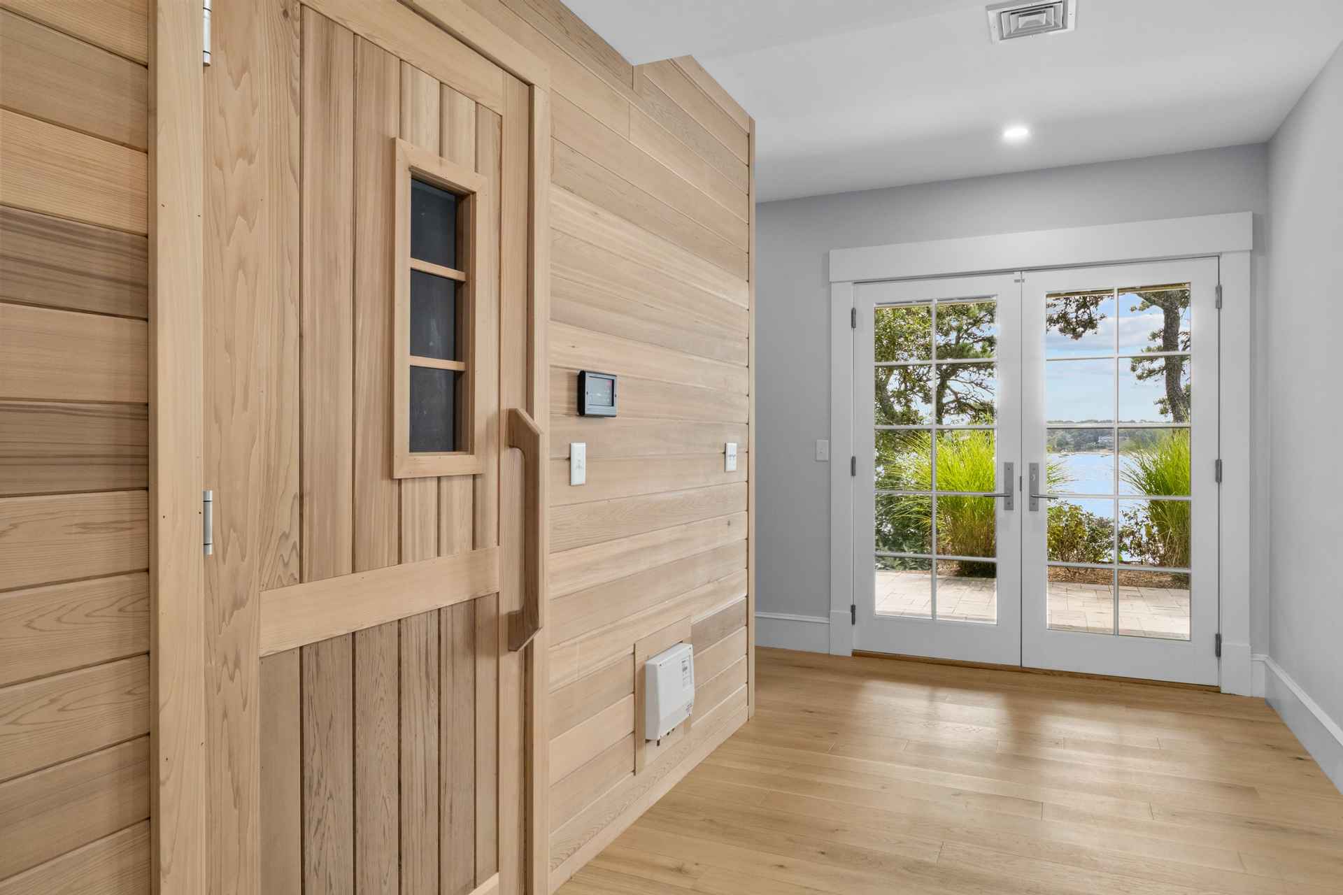 This interior shot showcases a hallway featuring a light wood sauna with a small window and a modern control panel. The hallway has light hardwood flooring and leads to a set of double doors with glass panes, offering a view of the outdoors. The walls are painted in a neutral gray tone, creating a bright and airy atmosphere.