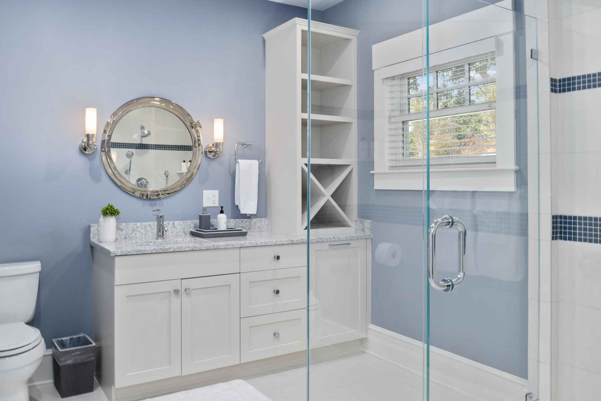 This is a well-lit bathroom featuring a vanity with white cabinets and a light-colored countertop, complemented by a round mirror and sconce lighting. A tall, narrow storage unit stands next to the vanity, and a glass shower enclosure is visible on the right. The walls are painted a soft blue, creating a serene and clean atmosphere.