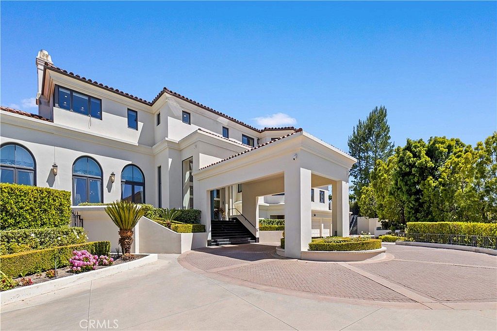 This grand estate features a prominent porte-cochère leading to a set of stairs at the main entrance, showcasing a sophisticated Mediterranean-inspired architectural style. The exterior is finished in a smooth, light-colored stucco, complemented by arched windows and a multi-level roofline. A circular driveway paved with patterned stone provides an elegant approach, framed by manicured hedges and mature trees under a clear blue sky.