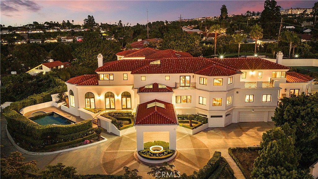 This expansive aerial view captures a grand Mediterranean-style estate at dusk, featuring a prominent circular motor court with a central fountain and a covered porte-cochère. The multi-level residence showcases a classic red-tiled roof, white stucco walls, and numerous windows, complemented by a private swimming pool and manicured landscaping. The elevated perspective highlights the property's impressive scale and its integration into a lush, hilly residential neighborhood.