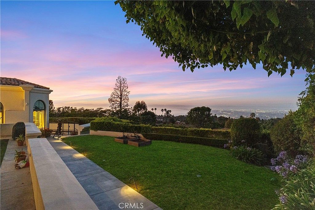 This elevated backyard offers a sweeping, panoramic view of the city lights and coastline under a vibrant sunset sky. The space features a manicured lawn, a stone-paved walkway, and raised garden beds, all framed by lush foliage and a large tree canopy. The perspective captures the serene transition from the home's architecture to the expansive horizon beyond.