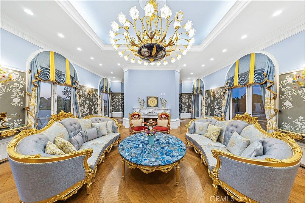 This formal living room showcases an opulent, neoclassical design with light blue walls, intricate crown molding, and a large, ornate gold chandelier as the centerpiece. The space is furnished with two matching curved sofas featuring light blue upholstery and gold frames, arranged around a striking circular blue agate coffee table. A fireplace with a detailed stone surround serves as a focal point at the far end of the room, flanked by two red-upholstered accent chairs, all set against a backdrop of patterned wallpaper and tall windows draped in elegant blue and gold curtains.