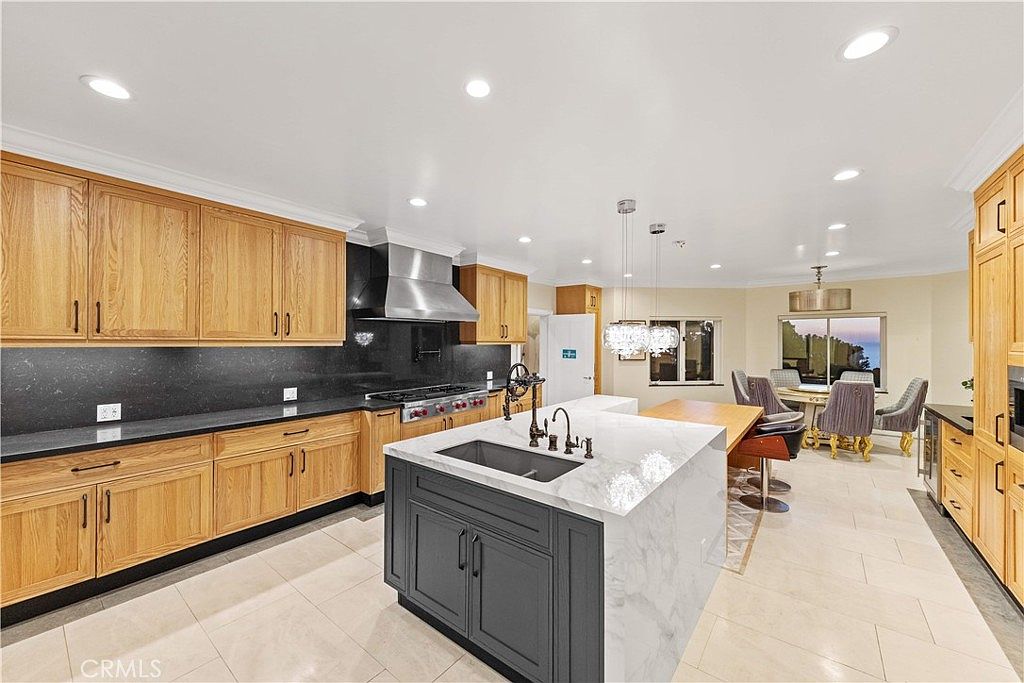 This spacious, open-concept kitchen features a large central island with a stone countertop and a dark-toned base, complemented by light wood cabinetry and dark stone countertops along the perimeter. A professional-style range with a stainless steel hood serves as a focal point, while a dining area with a round table and upholstered chairs is visible in the background. The room is brightly lit by recessed ceiling lights and decorative pendant fixtures, creating an inviting and functional culinary space.