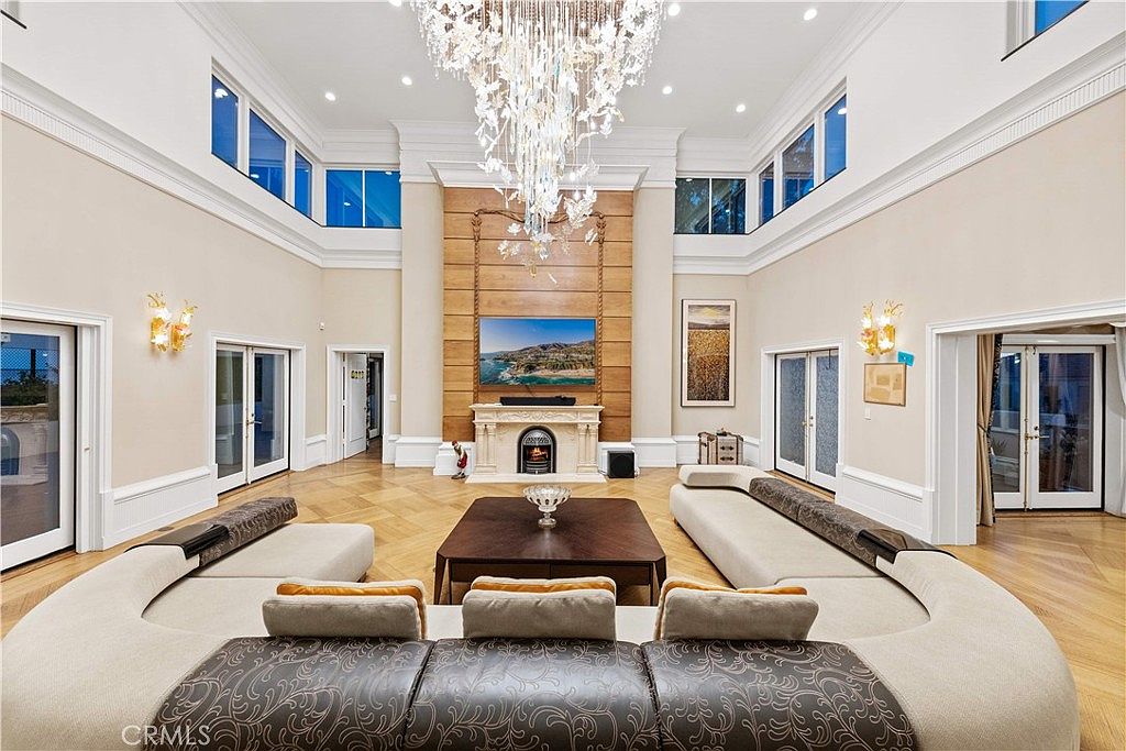 This expansive, double-height living room features a grand crystal chandelier as its centerpiece, hanging above a large, dark wood coffee table and a curved, neutral-toned sectional sofa. The focal point is a stone-clad chimney breast with a built-in television and a traditional fireplace, flanked by wood-paneled accents. The room is bathed in natural light from multiple tall windows, and the floor is finished with intricate, light-toned wood parquet.