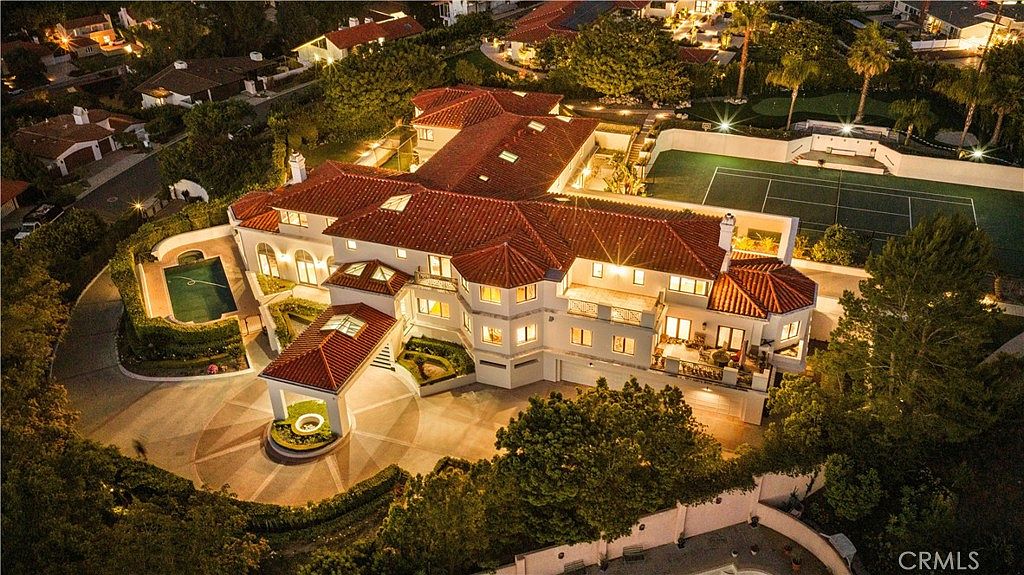 This high-angle aerial view captures a sprawling, multi-level luxury estate illuminated at night. The property features a prominent circular driveway with a covered porte-cochère, a private tennis court, and a swimming pool nestled within lush landscaping. The architecture showcases a classic Mediterranean style with red-tiled roofs and a complex, expansive footprint.