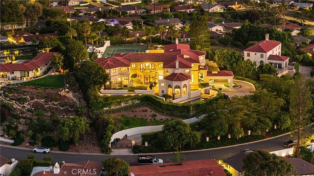 This high-angle aerial view captures a sprawling Mediterranean-style estate illuminated at dusk, showcasing its complex roofline with red clay tiles and warm interior lighting. The property features a private tennis court, a circular driveway, and lush landscaping that cascades down the hillside. The perspective emphasizes the grand scale of the residence and its prominent position within a quiet, upscale residential neighborhood.