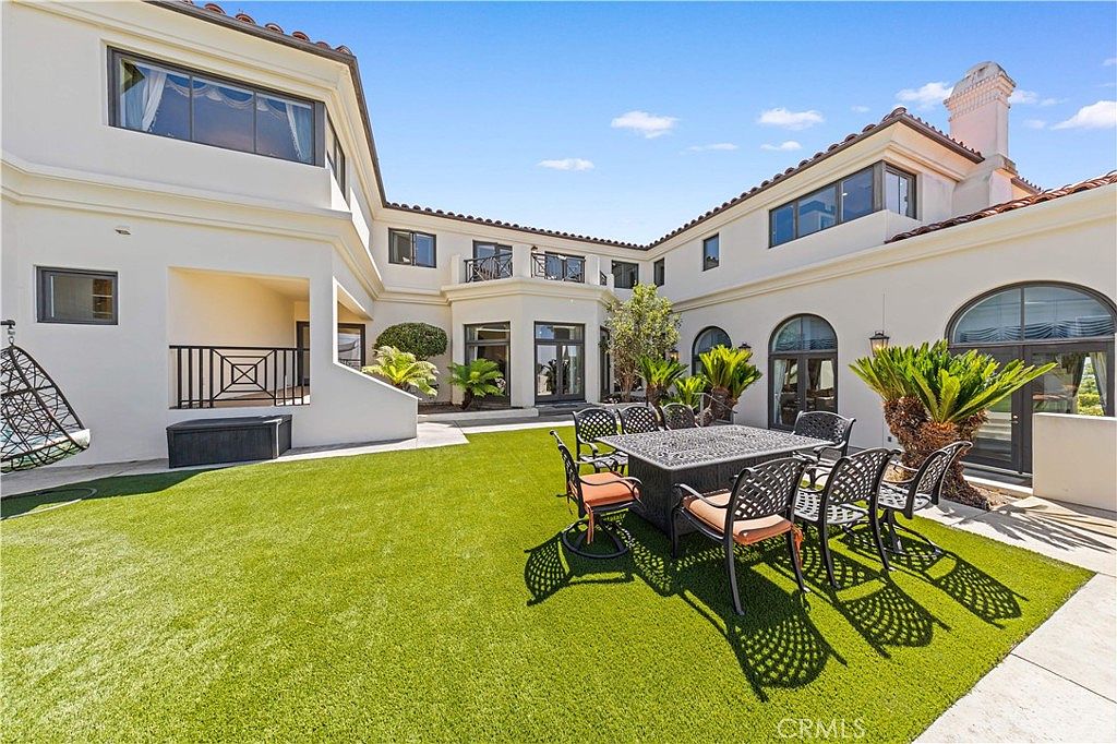 This image captures a spacious, U-shaped courtyard featuring a well-manicured artificial turf lawn and a dining area with a metal table and chairs. The exterior of the home is finished in a light-colored stucco with arched windows and doors, creating a Mediterranean-inspired aesthetic. A hanging chair sits to the left, while lush potted plants and mature landscaping add greenery to the bright, sunny outdoor space.