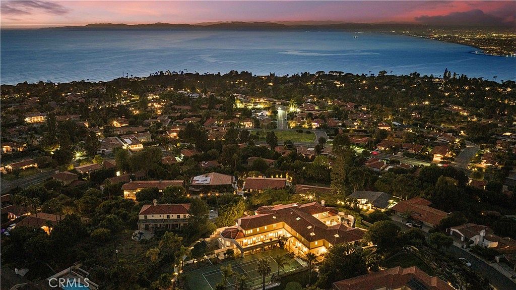 This high-angle aerial shot captures a sprawling luxury estate at twilight, set against a panoramic backdrop of the ocean and a distant coastline. The property features a large, multi-winged residence with a lit tennis court in the foreground, surrounded by lush landscaping and mature trees. The scene conveys a sense of exclusivity and prestige, highlighting the home's prime location within a quiet, upscale residential neighborhood.