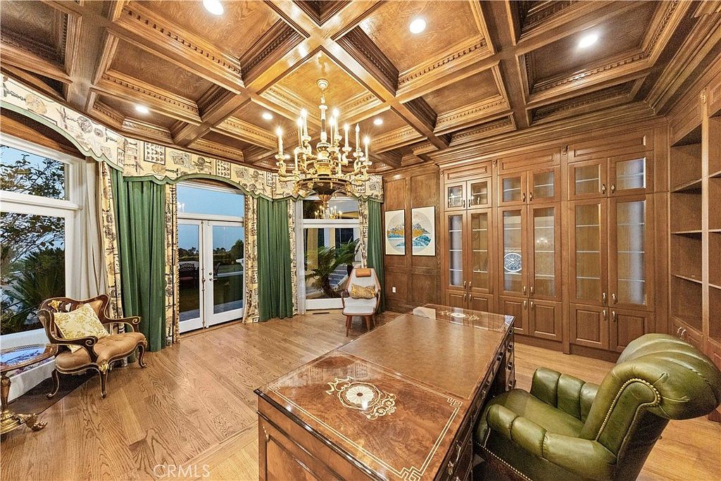 This sophisticated home office features extensive custom wood cabinetry and shelving that spans the wall, complemented by a coffered ceiling with recessed lighting and a central chandelier. A large, polished wood desk sits in the foreground, paired with a green leather armchair, while a secondary seating area is positioned near the tall windows draped in green curtains. The room exudes a traditional, executive atmosphere with its rich wood tones and classic architectural detailing.