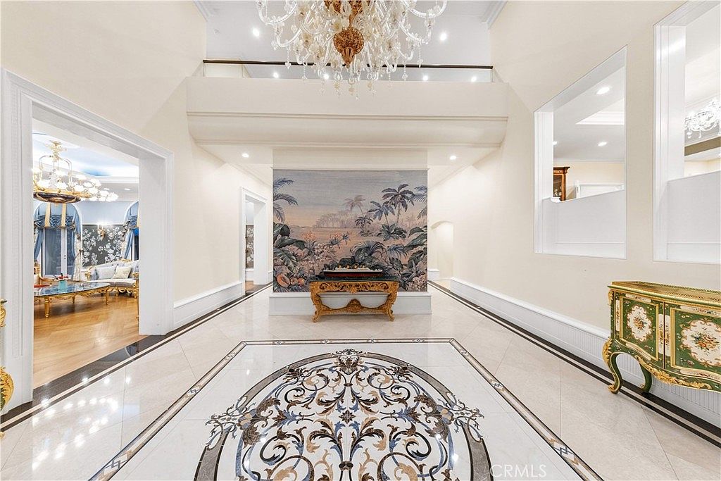 This grand foyer features a dramatic double-height ceiling with an ornate crystal chandelier and a central wall adorned with a large, detailed mural. The polished stone flooring showcases an intricate, circular inlay pattern, while a decorative console table sits against the mural wall. The perspective looks directly through the open entryway into an adjacent room, emphasizing the expansive and opulent scale of the home's interior design.