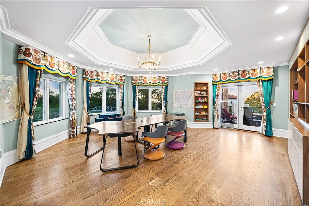 This spacious home office or study features a large, central wooden desk surrounded by modern chairs, set beneath an elegant octagonal tray ceiling with a central chandelier. The room is bathed in natural light from multiple windows adorned with decorative valances and curtains, and it includes built-in shelving units along the wall. The perspective is a wide-angle shot from the doorway, showcasing the polished wood flooring and the open, airy layout of the room.