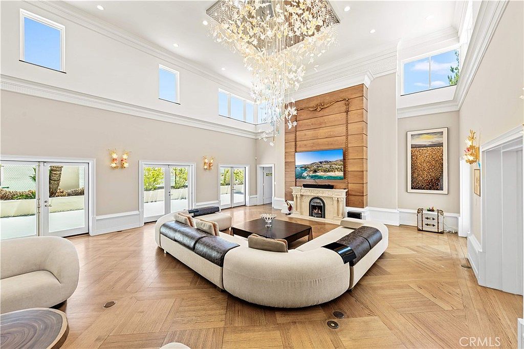 This expansive living room features a dramatic, multi-tiered crystal chandelier suspended from a high, coffered ceiling. A large, curved modular sofa centers the room, facing a stone fireplace with a tall, wood-paneled chimney breast and a mounted television. The space is brightened by multiple sets of French doors and large windows, all set against light-toned walls and warm-toned herringbone wood flooring.