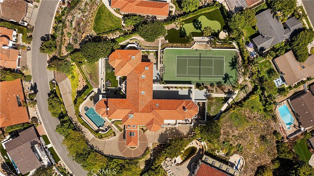 This high-angle aerial view captures a sprawling luxury estate featuring a large, multi-winged residence with a terracotta-tiled roof. The property is exceptionally well-appointed, boasting a private tennis court, a putting green, and a swimming pool nestled near the main structure. The surrounding landscape is lush with mature trees and manicured grounds, emphasizing the expansive and private nature of this residential compound.