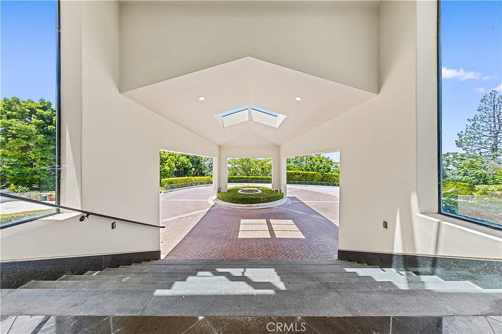This grand entryway features a set of wide, dark stone steps leading down from an interior space toward a covered outdoor motor court. The structure is defined by a high, vaulted ceiling with skylights, flanked by large glass panels that provide an expansive view of the surrounding lush greenery. The circular driveway below is paved with patterned brick and centers on a manicured circular garden bed, creating a sophisticated and welcoming arrival experience.