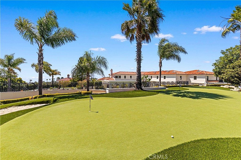 This expansive backyard features a professionally manicured putting green that serves as the focal point of the outdoor space. The property includes mature palm trees and a sprawling, single-story residence with a classic Mediterranean-inspired aesthetic, characterized by a low-pitched tiled roof and light-colored stucco walls. The scene is captured from a low angle, emphasizing the vastness of the lawn and the seamless integration of recreational amenities with the home's architecture.