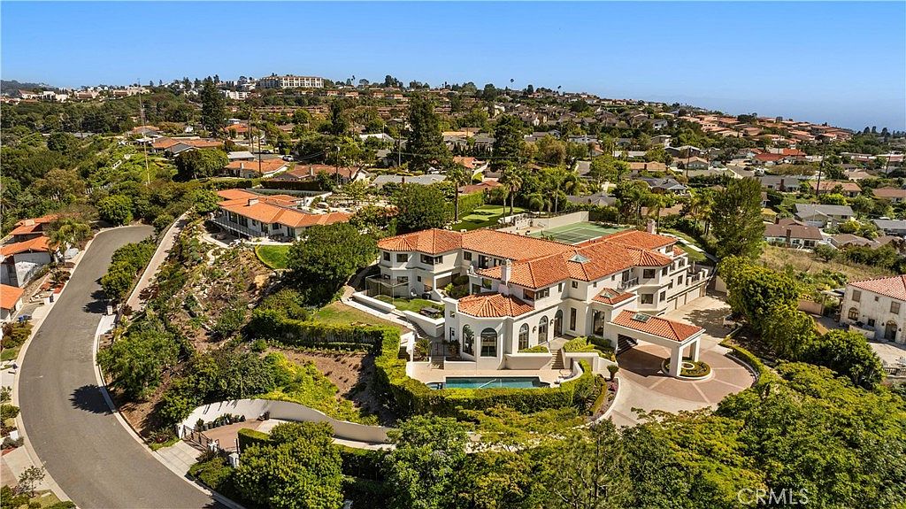 This high-angle aerial view captures a sprawling Mediterranean-style estate nestled in a lush, hilly landscape. The property features a multi-level residence with a terracotta tile roof, a private swimming pool, and a circular driveway, all surrounded by mature trees and manicured landscaping. The perspective highlights the expansive layout of the home and its integration into the surrounding residential neighborhood.