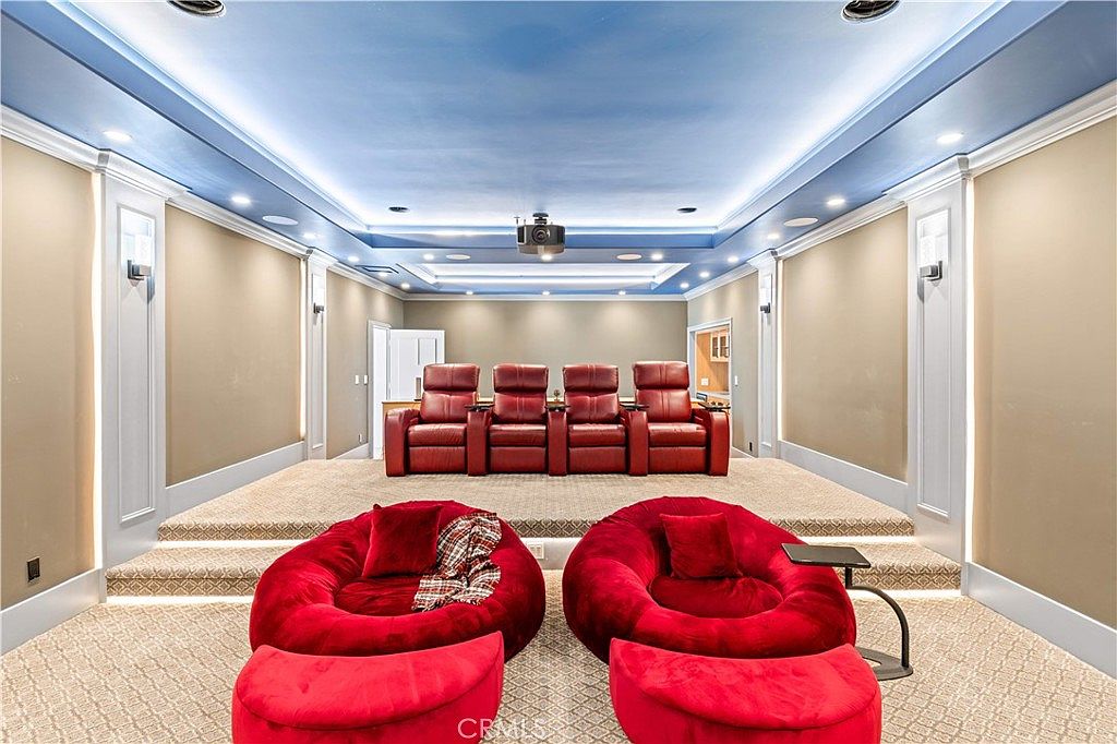This dedicated home theater room features a tiered layout with four plush red leather recliners on an elevated platform and two large, circular red lounge chairs in the foreground. The room is accented by recessed tray ceiling lighting and soft wall sconces, creating a cozy and immersive cinematic atmosphere. A projector is mounted to the ceiling, centered above the seating area, highlighting the space's functionality as a private entertainment hub.