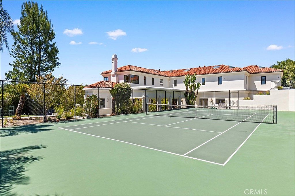 This image captures a private tennis court set against the backdrop of a large, multi-story Mediterranean-style residence with a terracotta tile roof. The court is enclosed by a black chain-link fence, and the surrounding landscape features mature trees and manicured shrubs. The perspective is from the court level, looking toward the rear of the home, emphasizing the expansive outdoor recreational space.