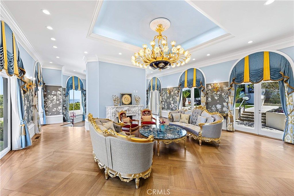 This elegant, formal living room features a sophisticated blue and gold color palette with ornate, traditional-style furniture and a grand crystal chandelier as the centerpiece. The space is defined by light wood flooring, intricate wall paneling with floral accents, and large arched windows that provide ample natural light and views of the outdoors. A central stone fireplace with a decorative mantel serves as a focal point, creating a luxurious and refined atmosphere.