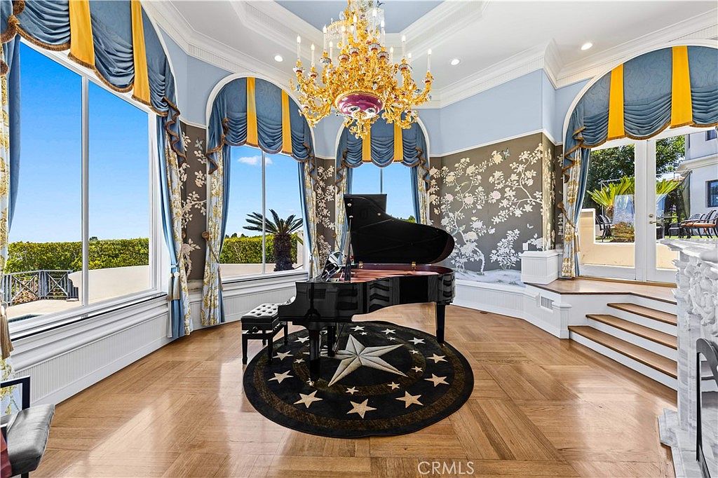 This elegant formal living space features a grand piano centered on a circular rug with a star motif, set against a backdrop of ornate floral wallpaper and tall windows draped in blue and gold fabric. A large, intricate gold chandelier hangs from the ceiling, casting light over the polished wood flooring. The room includes a raised platform area with French doors leading to an outdoor space, creating a sophisticated and airy atmosphere.