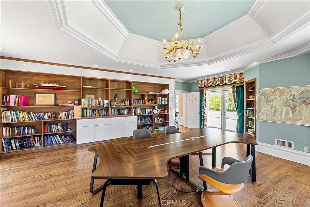 This spacious home office features a large, custom-shaped wooden desk centered in the room under an ornate chandelier hanging from a tray ceiling with a light blue accent. A wall of built-in wooden shelving provides extensive storage and display space, while a large world map hangs on the adjacent wall. The room is brightened by natural light streaming through glass doors that lead to an outdoor area, creating a professional yet inviting workspace.