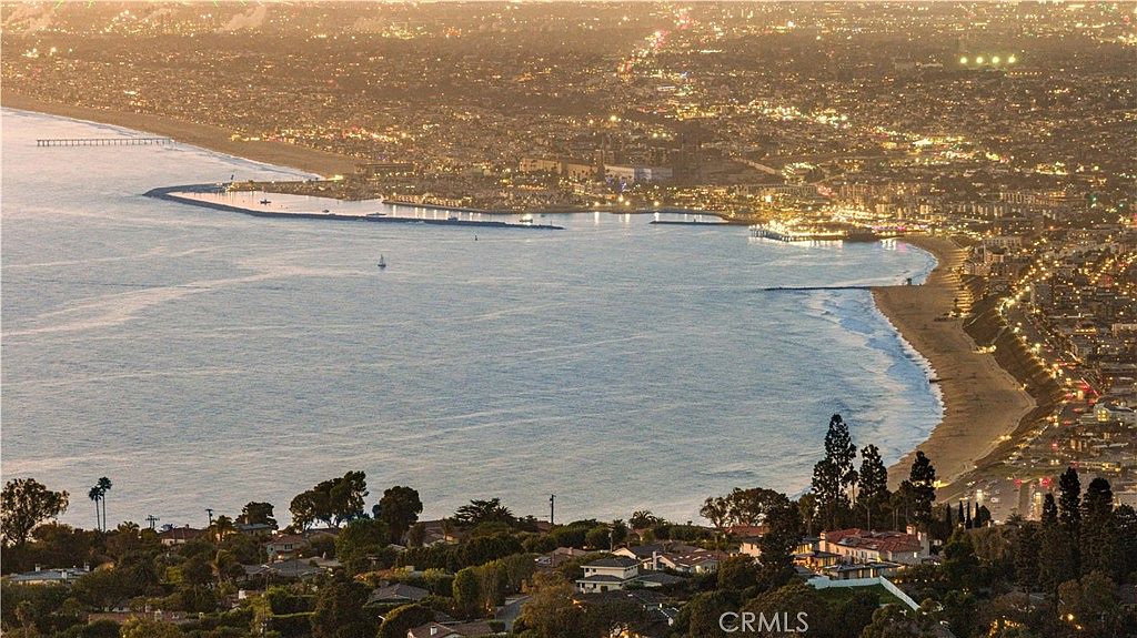 This high-angle aerial view captures a sprawling coastal landscape at twilight, showcasing a residential neighborhood nestled on a hillside overlooking the ocean. The scene features a sweeping coastline with a pier extending into the water, illuminated by the warm glow of city lights and street lamps. The perspective emphasizes the property's prime location, highlighting the expansive panoramic views of the bay and the surrounding urban development.