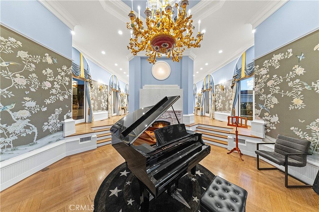 This elegant living room features a grand piano centered on a star-patterned rug, set against a backdrop of ornate floral wallpaper and light blue walls. The space is defined by a symmetrical layout with raised platforms on either side, tall windows draped in blue curtains, and a prominent, intricate gold chandelier hanging from the ceiling. A fireplace with a stone surround serves as a focal point, creating a sophisticated and formal atmosphere.