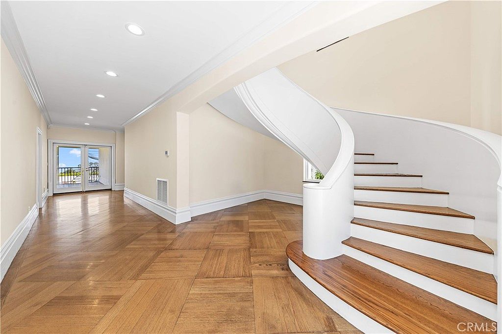 This elegant foyer features a prominent curved staircase with a white balustrade and rich, dark wood treads. The space is finished with light-toned walls and intricate parquet wood flooring, creating a bright and sophisticated atmosphere. A set of double glass doors at the end of the hallway provides natural light and a view of the outdoor balcony.