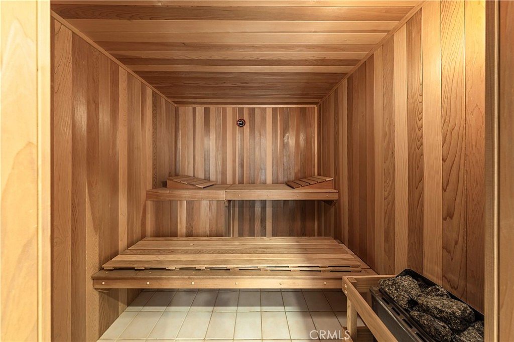 This image shows a traditional wood-paneled sauna featuring tiered benches and a dedicated heater unit filled with stones. The interior is lined with light-toned wood planks, creating a warm and inviting atmosphere, while the floor is finished with light-colored square tiles. The perspective is centered, looking directly into the sauna, highlighting the clean, well-maintained condition of the space.