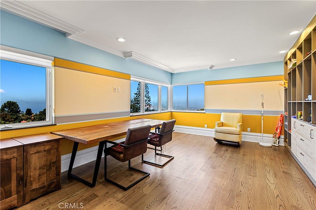 This bright and spacious home office features a long, natural-edge wood desk paired with two leather chairs, positioned to take advantage of the panoramic views through large windows. The walls are painted in a tri-color scheme of light blue, cream, and yellow, complemented by a large built-in shelving unit along the right wall. A comfortable yellow armchair sits in the corner, creating a cozy reading nook within this functional workspace.