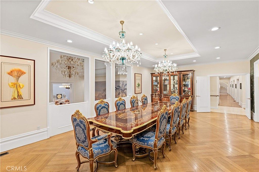 This formal dining room features a grand, long table surrounded by ornate, blue-upholstered chairs, set beneath two elaborate crystal chandeliers. The space is defined by its polished wood parquet flooring, a coffered ceiling, and a large, dark wood china cabinet against the back wall. A wide opening leads to a long hallway, while a decorative interior window provides a view into an adjacent room, creating an elegant and spacious atmosphere.