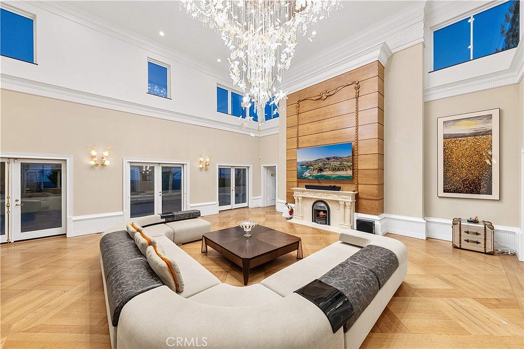 This expansive living room features high ceilings, light-toned walls, and intricate herringbone-patterned wood flooring. A large, curved sectional sofa centers the room around a dark wood coffee table, while a prominent fireplace with a decorative mantel and a tall wood-paneled surround serves as the focal point. The space is illuminated by a grand crystal chandelier and natural light streaming through multiple tall windows.