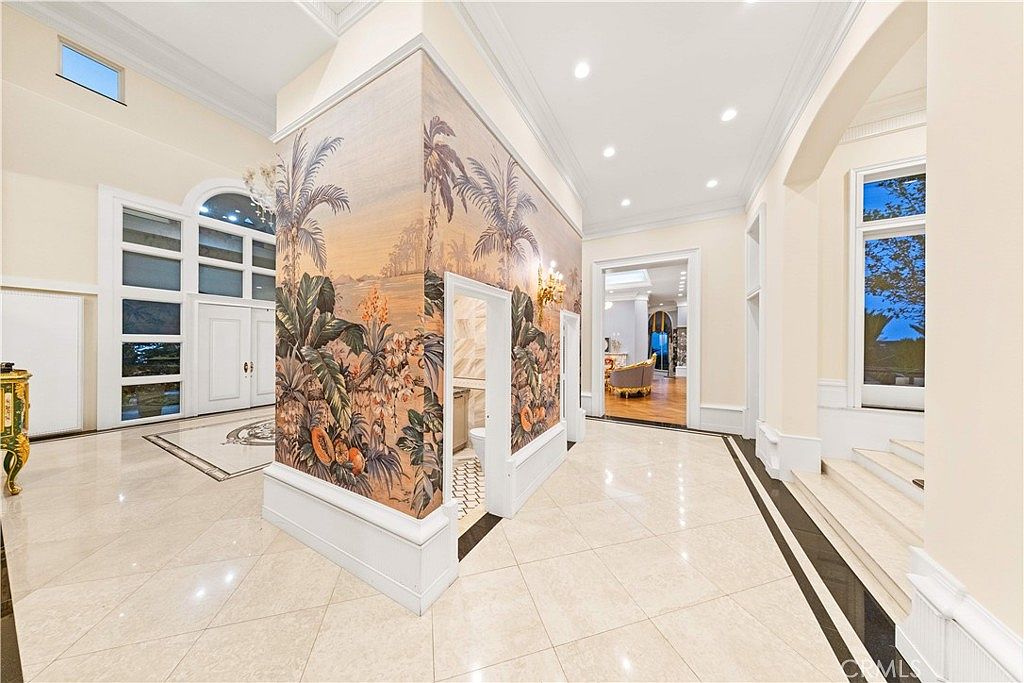 This grand foyer features high ceilings and polished light-colored tile flooring with a dark border inlay. A central structural column is adorned with a vibrant, tropical-themed mural, which also conceals a small powder room entrance. To the right, a set of stairs leads upward, while the open floor plan provides a clear line of sight into an adjacent living area with hardwood floors.