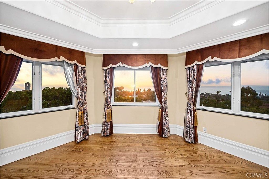 This bright, expansive room features a bay window configuration that offers panoramic ocean views. The space is framed by elegant valanced curtains and features light-toned wood flooring and a tray ceiling with recessed lighting. The neutral wall color and symmetrical layout create a sophisticated and airy atmosphere suitable for a formal living or sitting area.