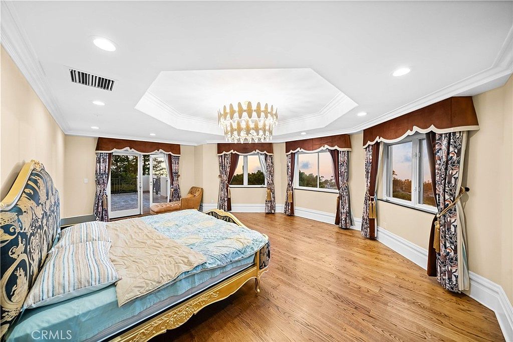This spacious primary bedroom features a tray ceiling with a central chandelier and warm-toned wood flooring. The room is furnished with a large bed featuring a decorative patterned headboard and offers access to an outdoor balcony through glass doors. Multiple windows with elegant drapery provide natural light and views of the surrounding area.