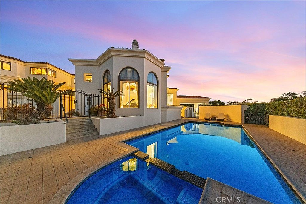 This image captures a luxurious backyard featuring a pristine swimming pool and an integrated spa at sunset. The home's exterior showcases elegant architectural details, including tall arched windows that reveal a glimpse of the interior, set against a vibrant, colorful sky. A paved patio area surrounds the water features, creating a sophisticated space for outdoor relaxation and entertaining.