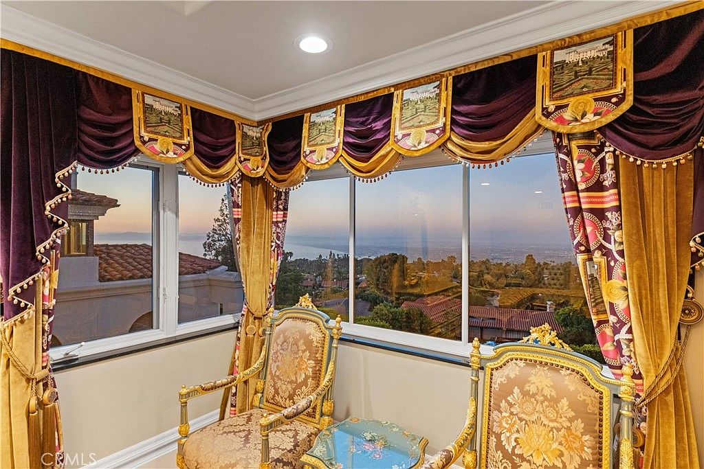 This elegant sunroom features ornate, heavy velvet drapery with gold trim and decorative valances framing large windows that offer a panoramic view of the coastline and surrounding landscape. Two matching antique-style chairs with floral upholstery and gold-toned frames are positioned around a small, decorative glass-topped table. The space exudes a formal, traditional aesthetic, emphasizing the dramatic views and luxurious window treatments.