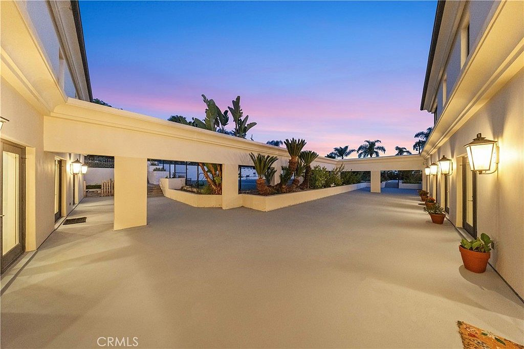 This expansive, covered outdoor patio area features a smooth, neutral-toned concrete floor and a long, sheltered walkway lined with wall-mounted lanterns. The space is framed by a clean, minimalist architectural structure with integrated planters containing lush greenery, creating a serene transition between the home's interior and the exterior. The perspective captures the long, open-air corridor at dusk, with the soft glow of the wall lights contrasting against the vibrant, colorful sunset sky.