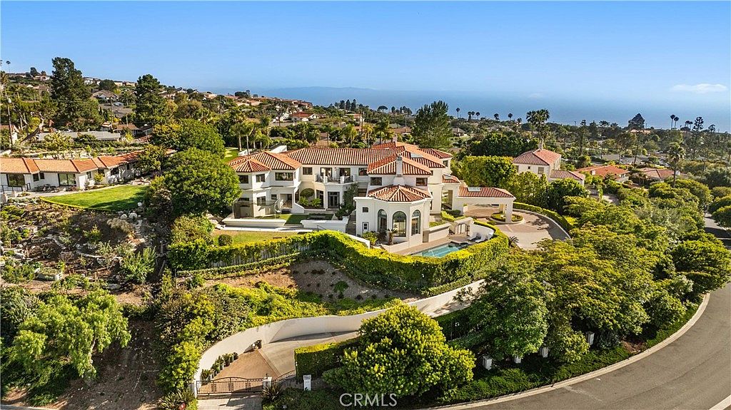 This high-angle aerial perspective captures a sprawling Mediterranean-style estate nestled on a hillside with expansive ocean views in the background. The property features a multi-level layout with terracotta-tiled roofs, a private swimming pool, and lush, manicured landscaping that cascades down the slope. A winding driveway leads to the residence, which is surrounded by mature trees and well-maintained greenery, creating a sense of privacy and luxury.