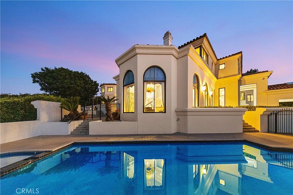 This rear exterior view captures a luxurious Mediterranean-style home at dusk, featuring a prominent curved architectural wing with tall windows. A pristine, rectangular swimming pool with a connected spa occupies the foreground, reflecting the warm glow emanating from the house's interior. The scene is framed by manicured landscaping, including a palm tree and neatly trimmed hedges, creating an inviting and upscale atmosphere.
