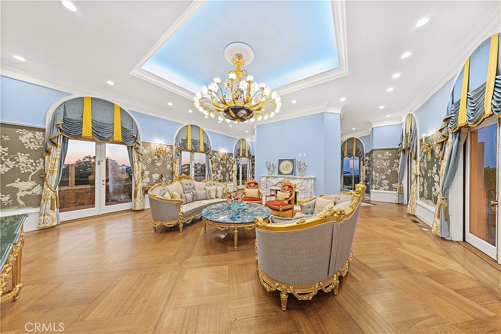 This expansive, formal living room features a sophisticated neoclassical design with light blue walls, intricate wall molding, and a coffered ceiling centered by a grand crystal chandelier. The space is furnished with ornate, gold-trimmed seating arranged around a circular stone-topped coffee table, all set upon polished light wood flooring. Large arched windows draped in blue and gold fabric provide ample natural light and views of the exterior, while a decorative fireplace serves as a focal point on the far wall.