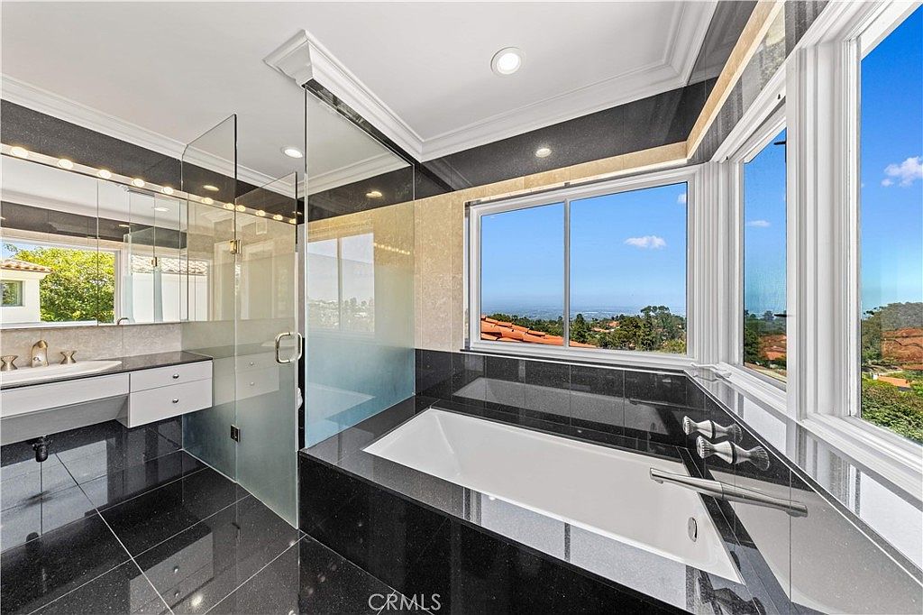 This luxurious primary bathroom features a deep soaking tub set against a backdrop of dark, polished stone surfaces and large windows that offer expansive views of the surrounding landscape. A glass-enclosed shower area is positioned to the left, complemented by a sleek, wall-mounted vanity with a minimalist sink. The high-contrast design creates a sophisticated and modern atmosphere, emphasizing the bright natural light streaming into the space.
