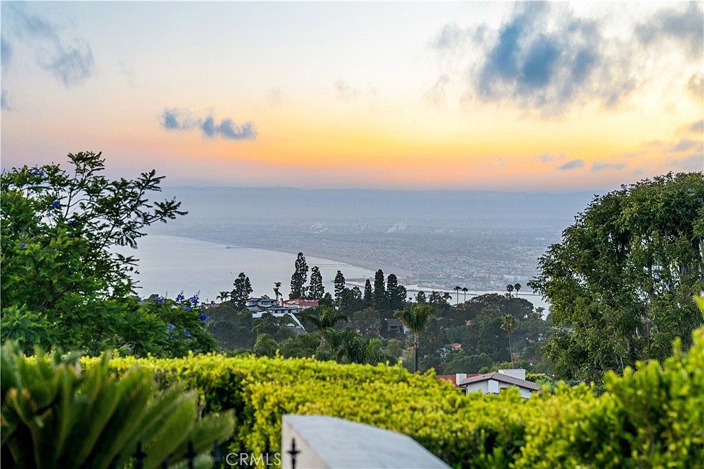 This elevated view captures a stunning coastal sunset overlooking a sprawling residential landscape and the Pacific Ocean. In the foreground, a manicured hedge frames the scene, while the mid-ground features lush greenery and rooftops nestled into the hillside. The horizon glows with soft orange and yellow hues, creating a serene and picturesque atmosphere ideal for a luxury property listing.