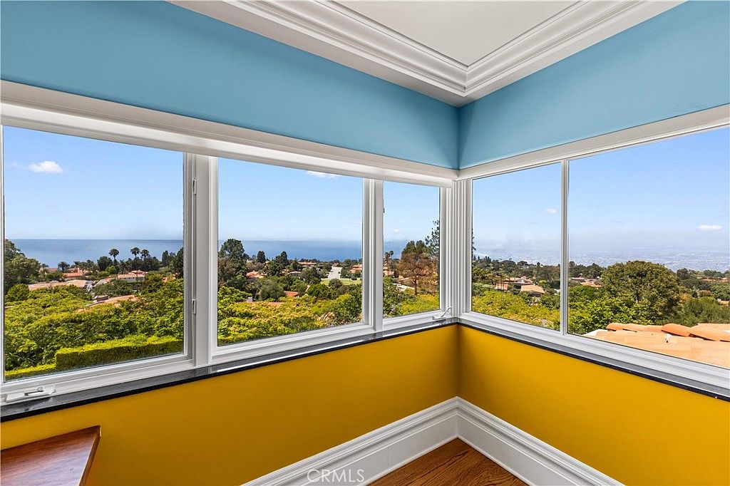 This bright sunroom features expansive windows that offer a panoramic view of the surrounding landscape and the distant ocean. The room is defined by its vibrant yellow lower walls, contrasting with a soft blue upper wall and crisp white crown molding. The warm wood flooring adds a natural touch to the space, creating an inviting atmosphere perfect for taking in the scenic vistas.
