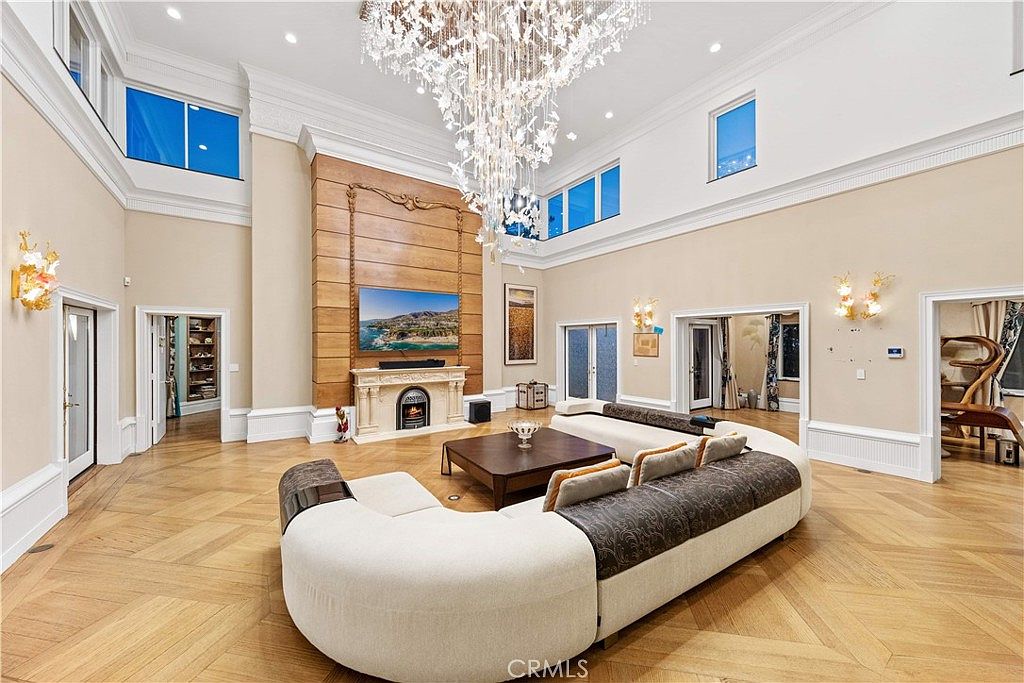 This expansive living room features a dramatic double-height ceiling anchored by a grand crystal chandelier and light-toned walls. A large, curved sectional sofa faces a central coffee table and a stone-mantel fireplace set against a prominent wood-paneled chimney breast. The space is finished with intricate herringbone-patterned wood flooring and multiple doorways leading to adjacent rooms, creating an open and airy atmosphere.