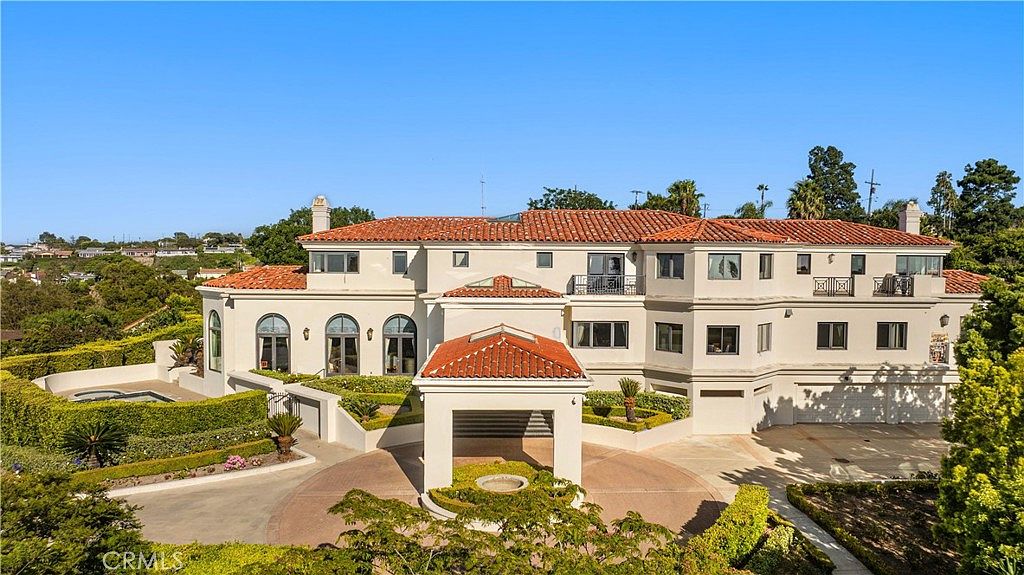 This grand, multi-story Mediterranean-style estate features a prominent porte-cochère at the entrance and a circular driveway. The exterior is finished in a light stucco with a classic red-tiled roof, complemented by manicured hedges and a small pool area to the left. The elevated perspective captures the expansive scale of the residence and its integration into the surrounding landscape.
