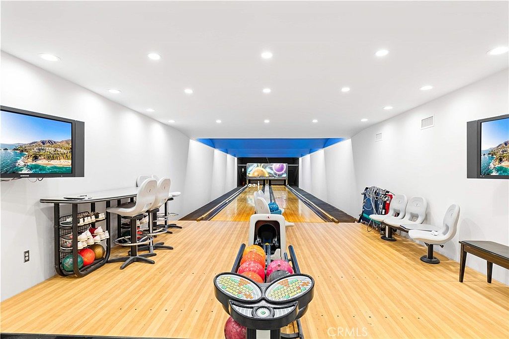 This image showcases a private, two-lane home bowling alley featuring a polished wood lane and a ball return mechanism in the foreground. The room is equipped with white molded seating, a high-top table with stools, and wall-mounted televisions on either side, creating a dedicated entertainment space. The perspective is centered down the length of the alley, highlighting the sleek, modern design and recessed ceiling lighting.